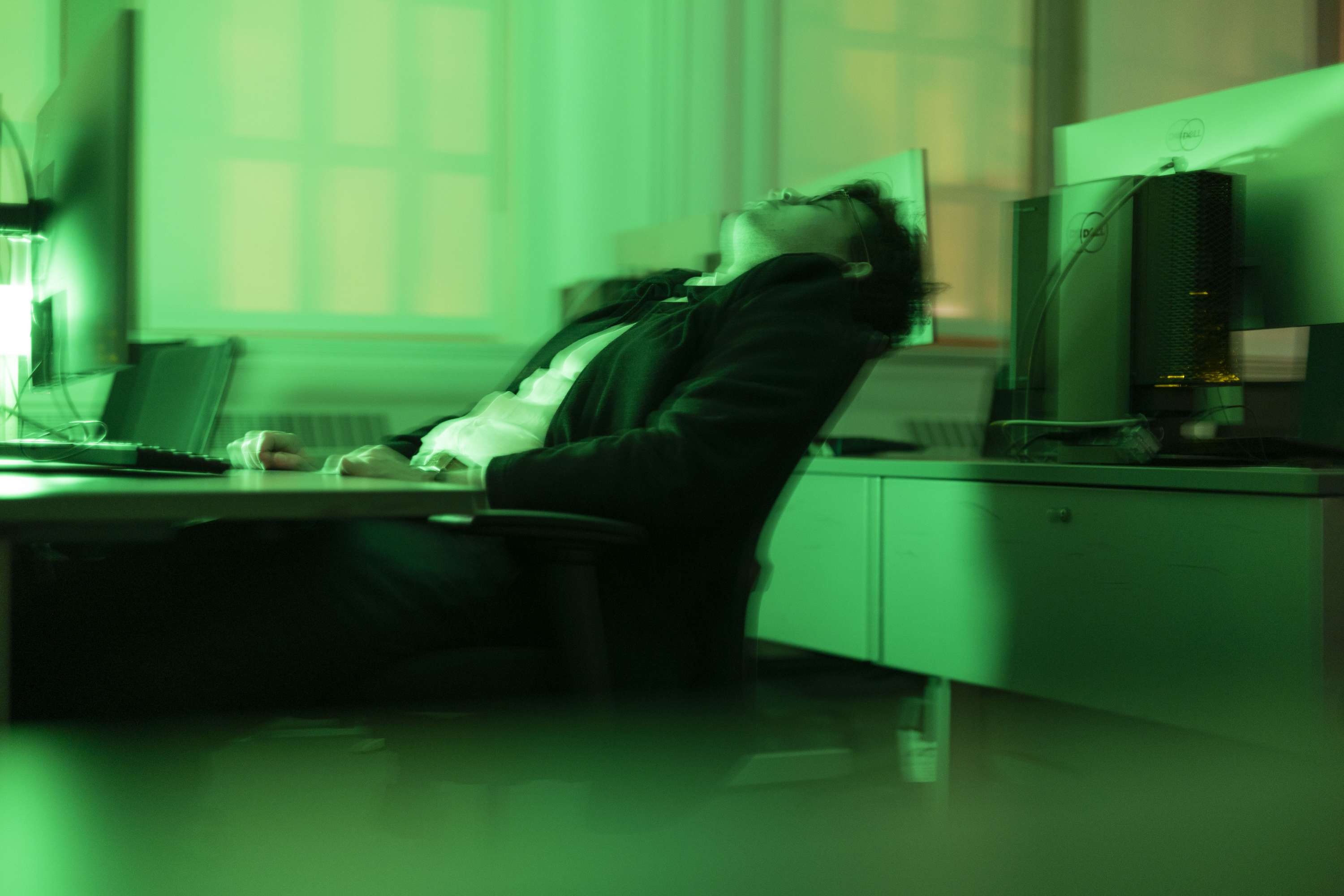 A person leaning back in a chair, surrounded by computers and green light.