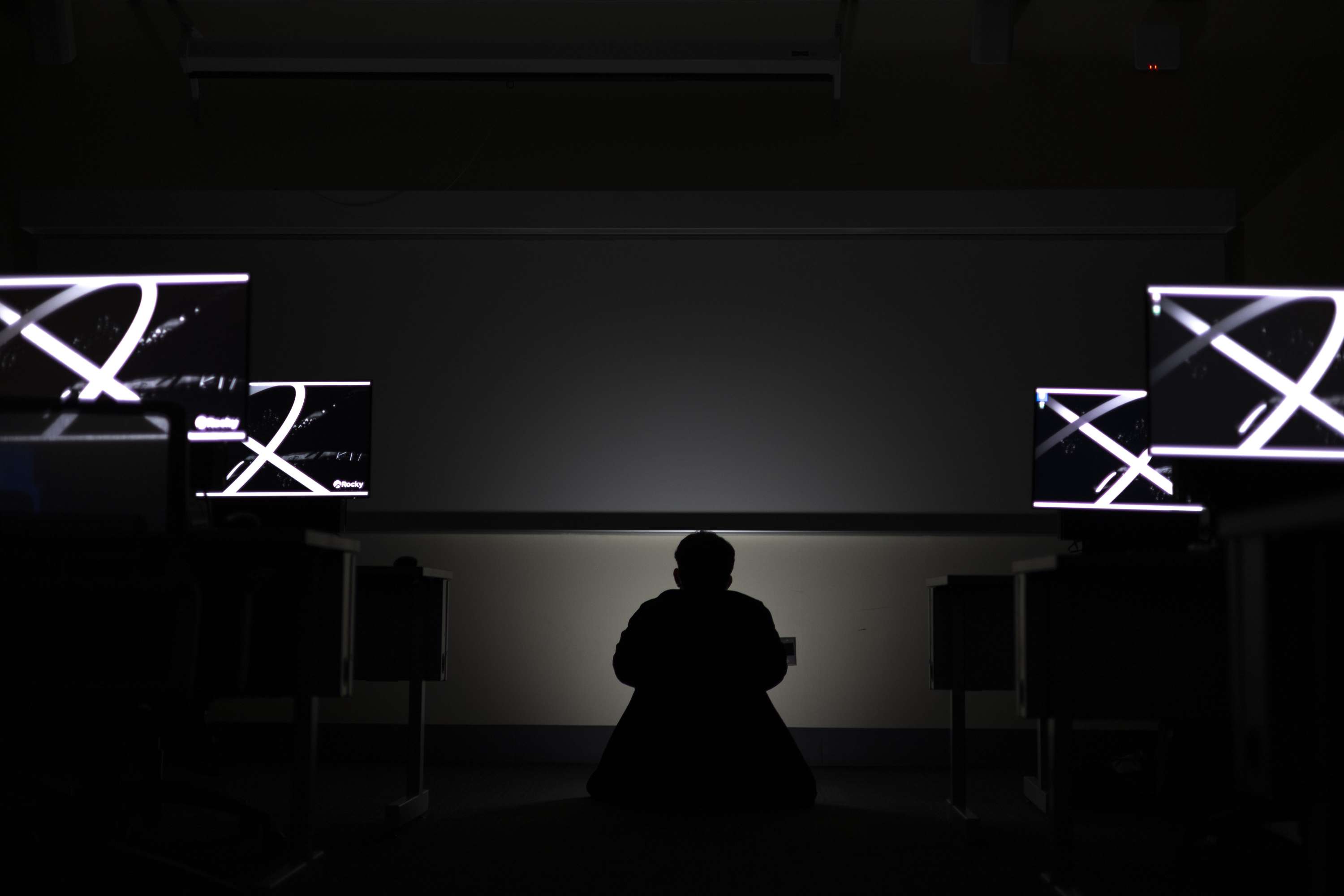 A seated silhouette at the center of a dark room, flanked by illuminated monitors.