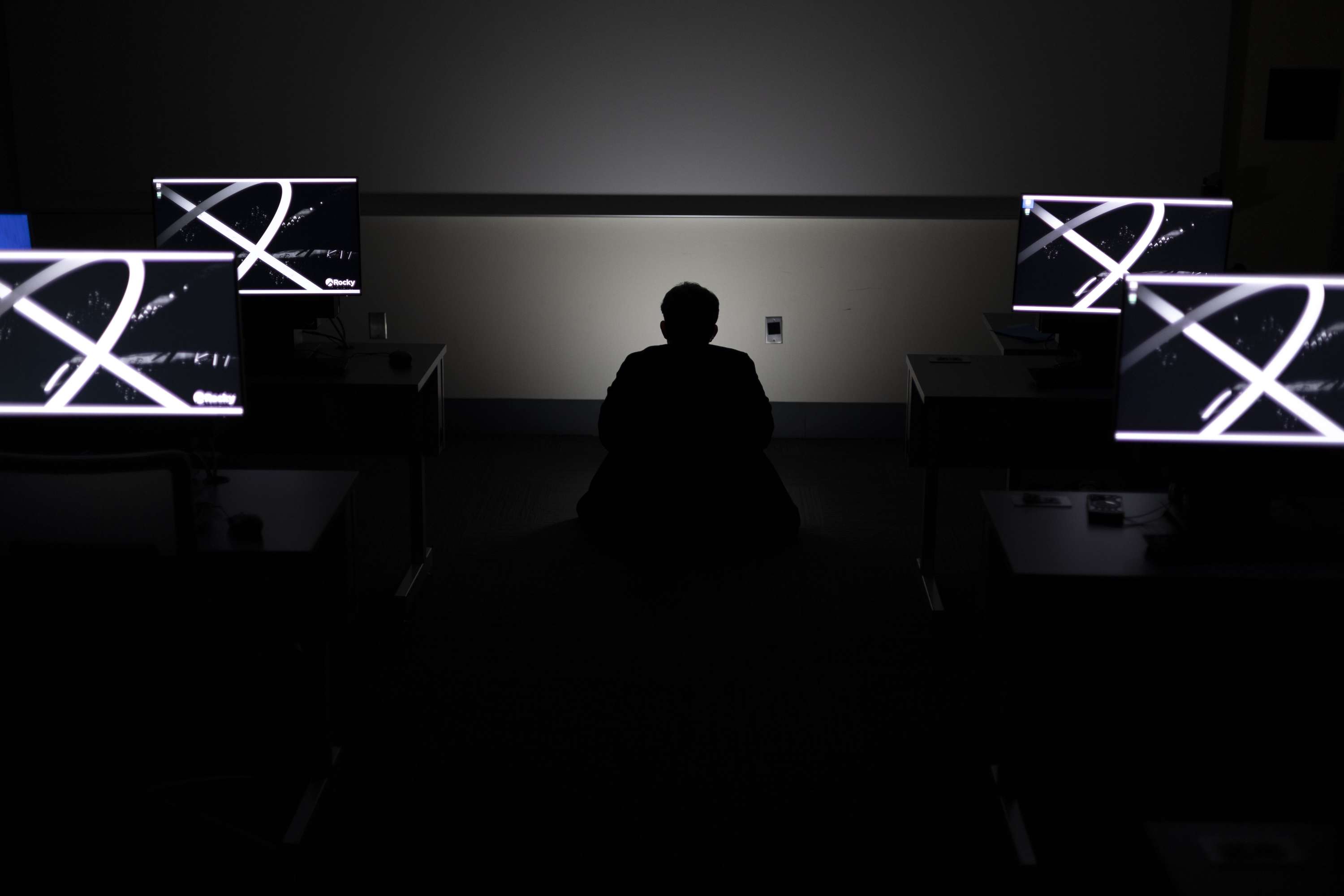 A seated figure between two symmetrical rows of bright monitors in a dark computer room.