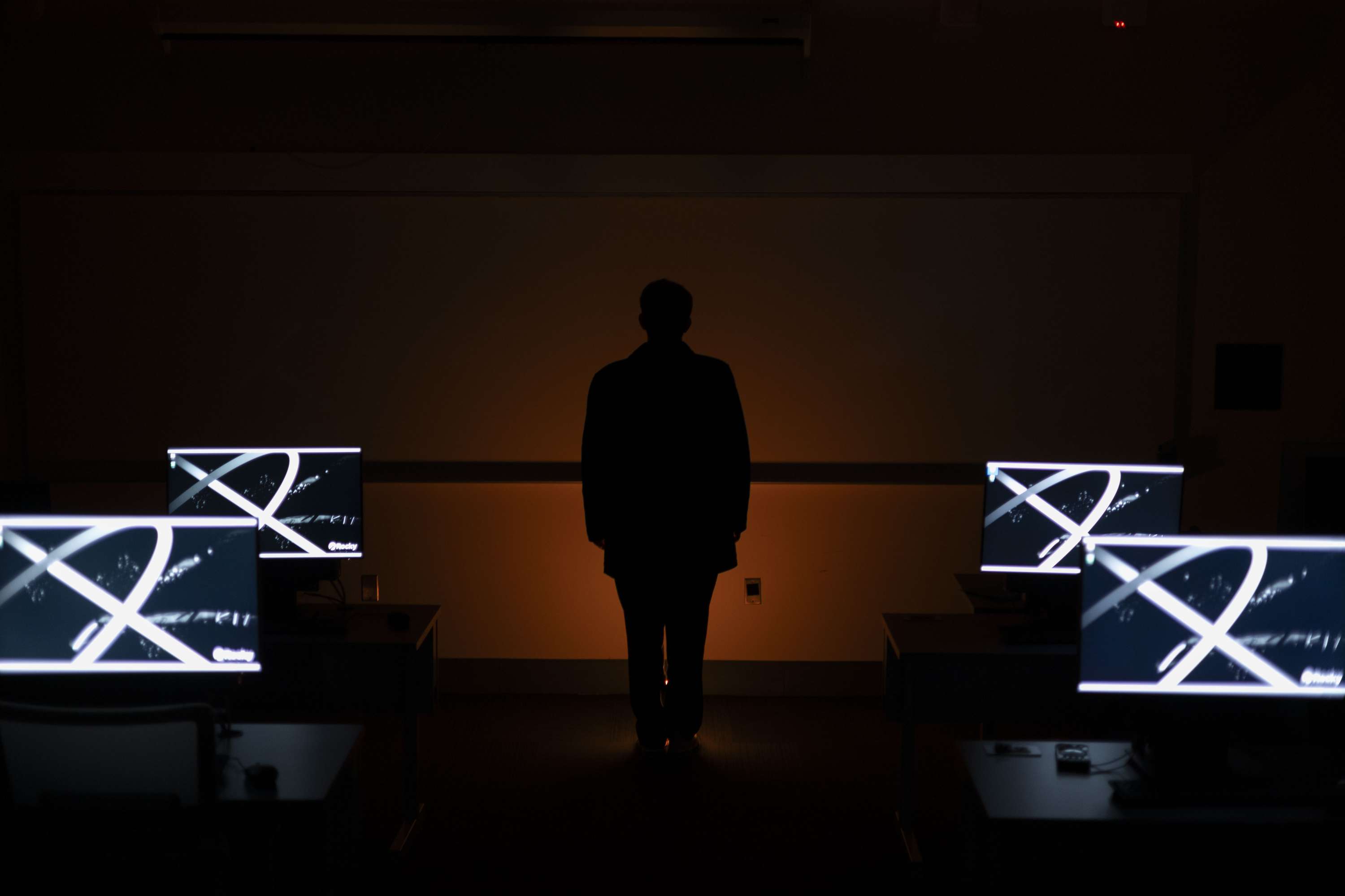 A standing silhouette against a warm orange-lit wall, surrounded by illuminated monitors.